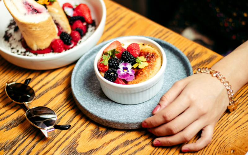 Vibrant crème brûlée with berries and flowers on table, next to berry-topped toast, hand with bracelet, evoking French dessert charm.