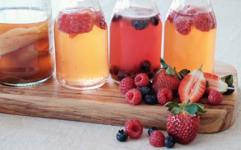 7 Assortment of colorful kombucha bottles topped with berries and strawberries on wooden board, highlighting fermented drink popularity.